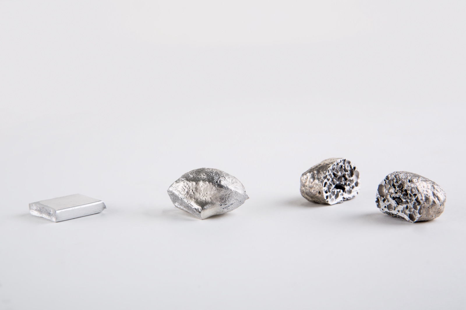 Vier Stadien nebeneinander: eine flache Vormaterialplatte aus Aluminium, ein teilweise aufgeschäumtes Stück und zwei aufgeschäumte Körper, deren angeschnittene Flächen die Zellen im Inneren zeigen.