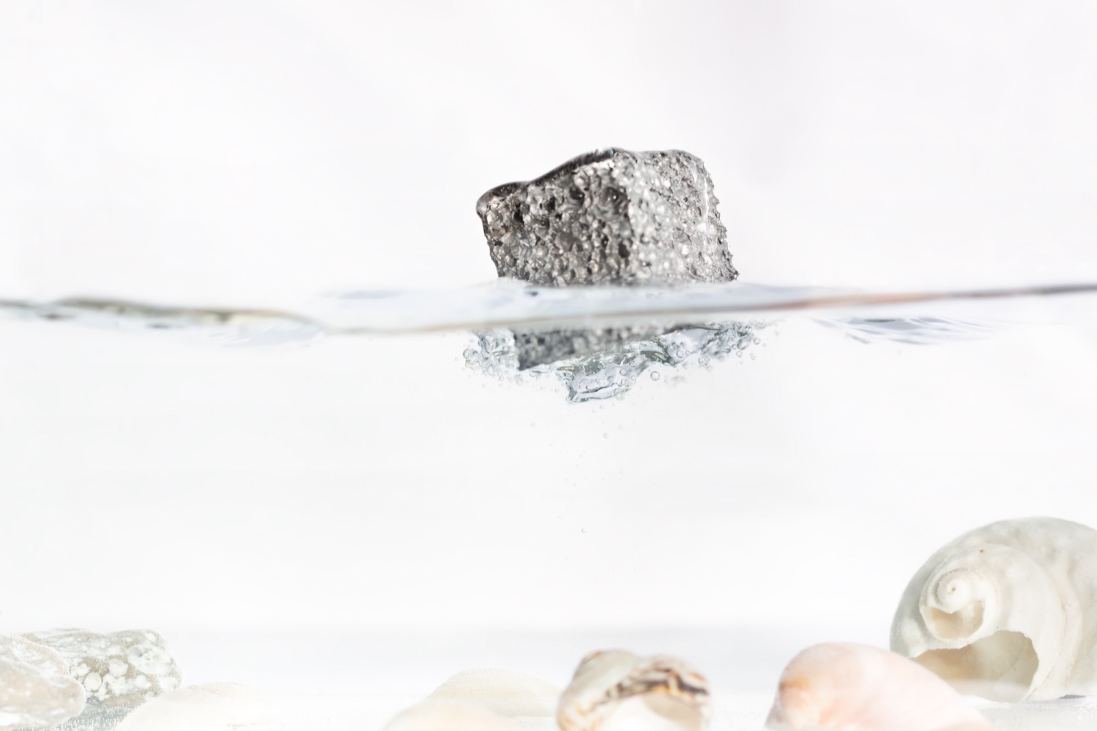 Ein Stück Aluminiumschaum liegt ruhig an der Wasseroberfläche und ragt zum größten Teil aus dem Wasser heraus; darunter steigen einzelne Luftblasen auf, am Beckenboden liegen Muscheln.