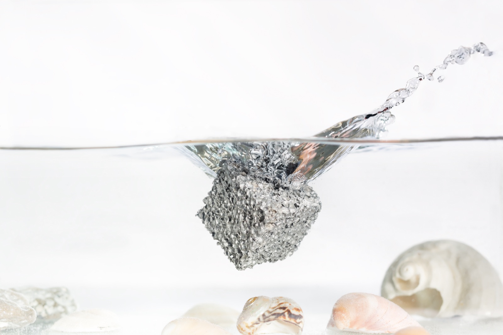 Ein Würfel aus Aluminiumschaum an der Wasseroberfläche, halb eingetaucht, mit aufspritzendem Wasser; am Boden des Beckens liegen Muscheln.