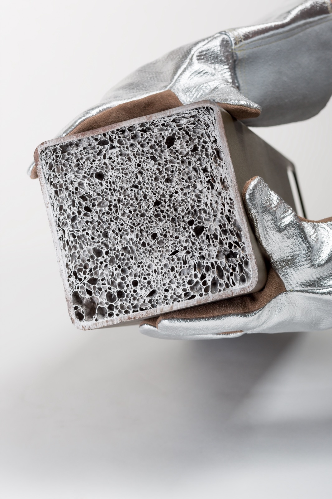 Eine behandschuhte Hand hält ein quadratisches Hohlprofil mit der Schnittfläche zum Betrachter; der Hohlraum ist vollständig mit Aluminiumschaum gefüllt, der bis an die Profilwände reicht.