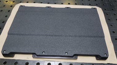 Dunkelgrau beschichtete Schutzplatte mit rauer Oberfläche, an der unteren Kante eine angeschraubte Halteleiste, liegend auf einem Lochraster-Tisch.