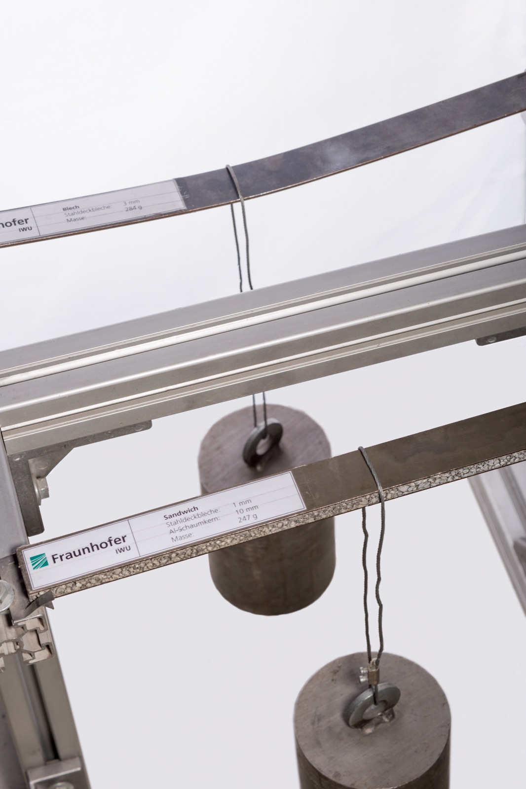 Ein Demonstrationsaufbau des Fraunhofer IWU mit zwei waagerecht eingespannten Proben, jede mit einem angehängten Gewicht: oben ein Stahlblech, das sich sichtbar durchbiegt, darunter ein Sandwich mit Stahldecklagen und Aluminiumschaumkern, das gerade bleibt. An beiden Proben hängen beschriftete Schilder.