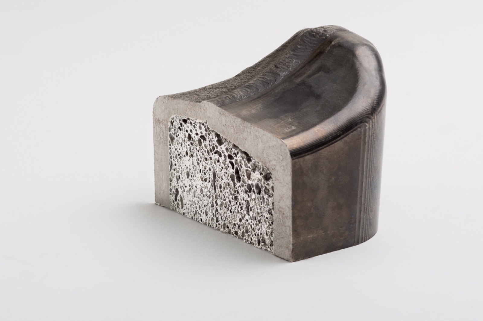 Ein Formteil mit sattelförmig gewölbter Oberseite und angeschnittener Stirnfläche, an der der Aluminiumschaumkern zwischen den Decklagen sichtbar wird; die gewölbte Fläche zeigt Anlauffarben.