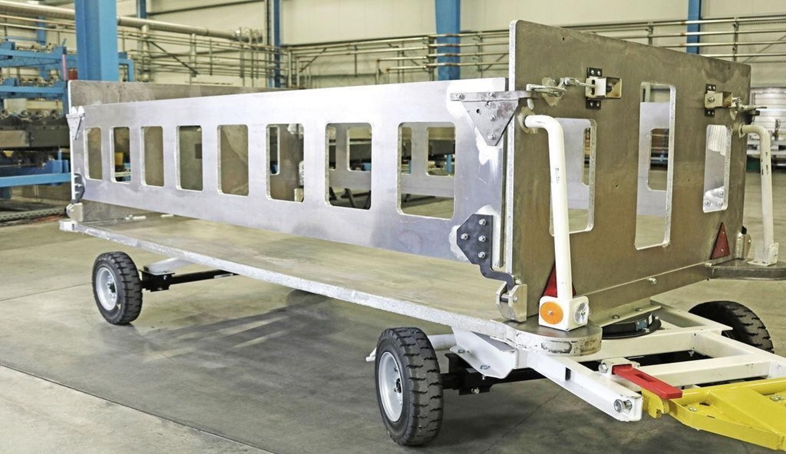 Ein Gepäckwagen in einer Fertigungshalle: Aufbau und Bordwände bestehen aus hellen Aluminium-Sandwichplatten mit ausgeschnittenen Fenstern, montiert auf einem Fahrgestell mit vier Rädern und Deichsel.