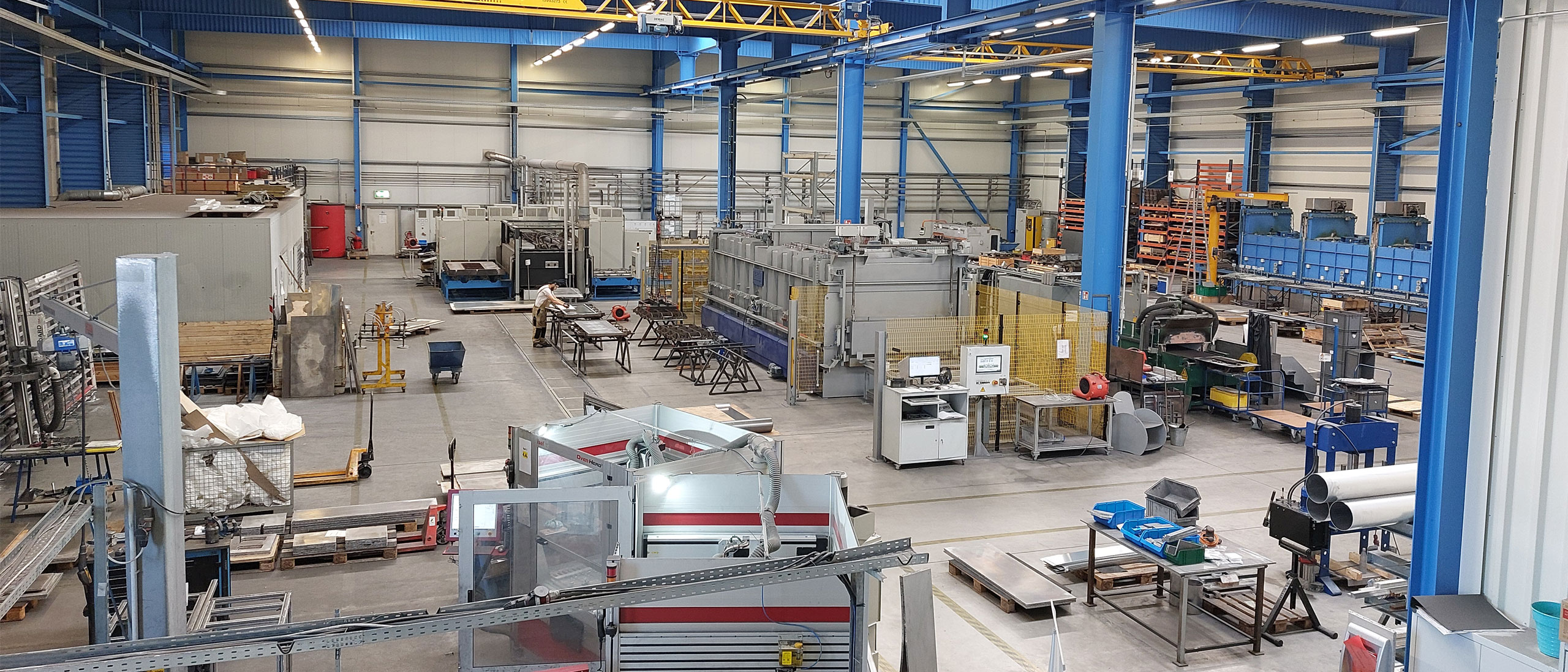Blick von einer Galerie in eine Fertigungshalle mit blauer Stahlkonstruktion und gelben Hallenkränen: in der Mitte eine lange graue Anlage hinter gelbem Schutzgitter, links ein Mitarbeiter an einem Arbeitstisch, im Vordergrund Maschinen und Paletten.