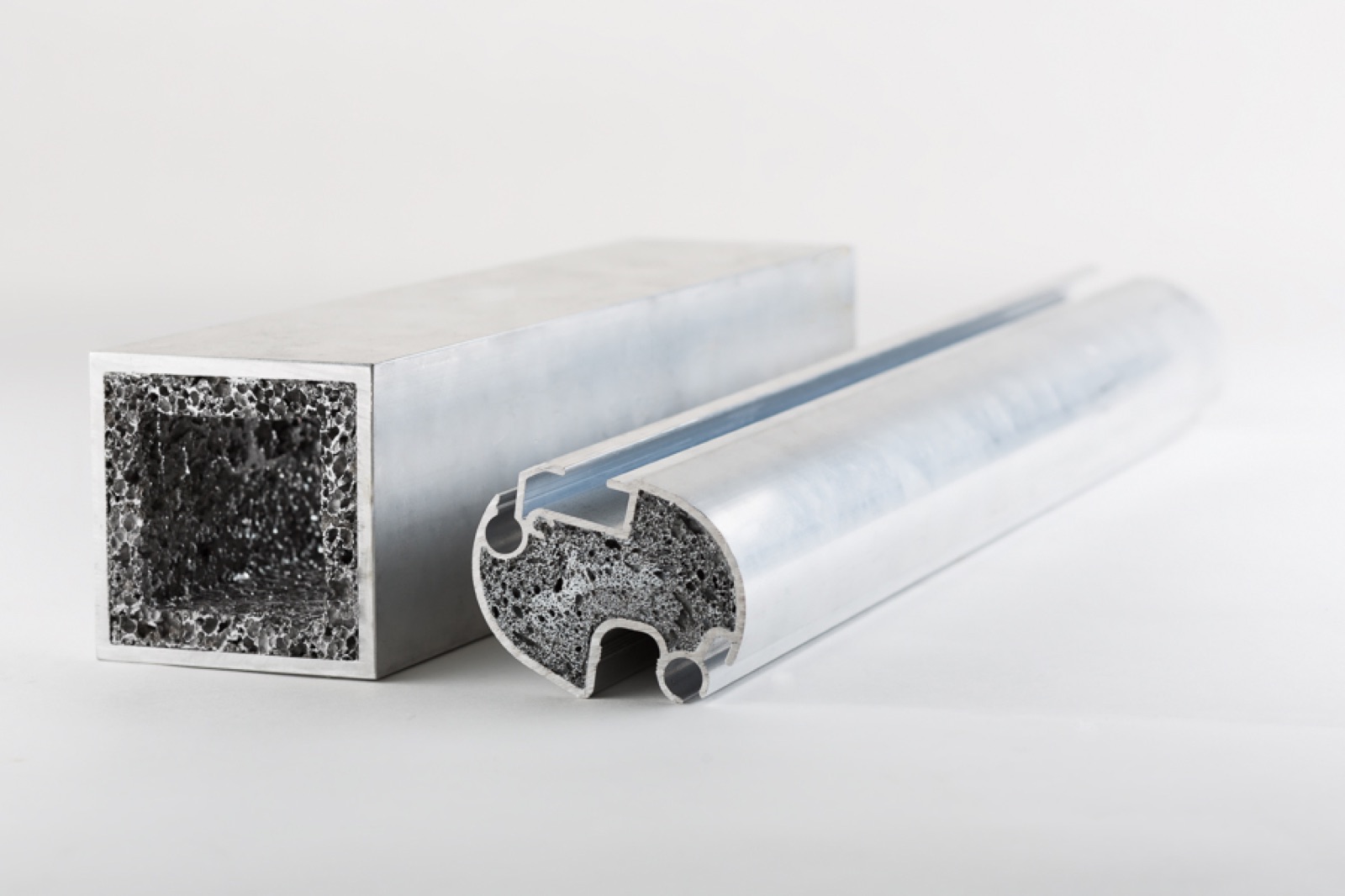 Zwei ausgeschäumte Aluminiumprofile: links ein Vierkantrohr, dessen Schnittfläche eine umlaufende Schaumschicht an den Innenwänden zeigt, rechts ein Rundprofil mit Nuten, in dessen Kammer der Schaum sitzt.