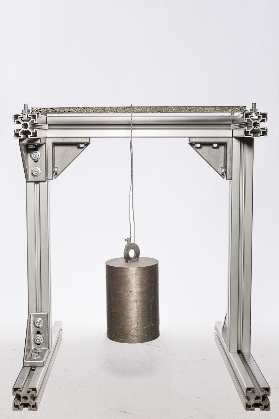 Ein Prüfgestell aus Aluminiumprofilen: Oben liegt waagerecht eine Sandwichprobe, an deren Mitte ein zylindrisches Stahlgewicht an einem Drahtseil hängt. Die Probe bleibt gerade.