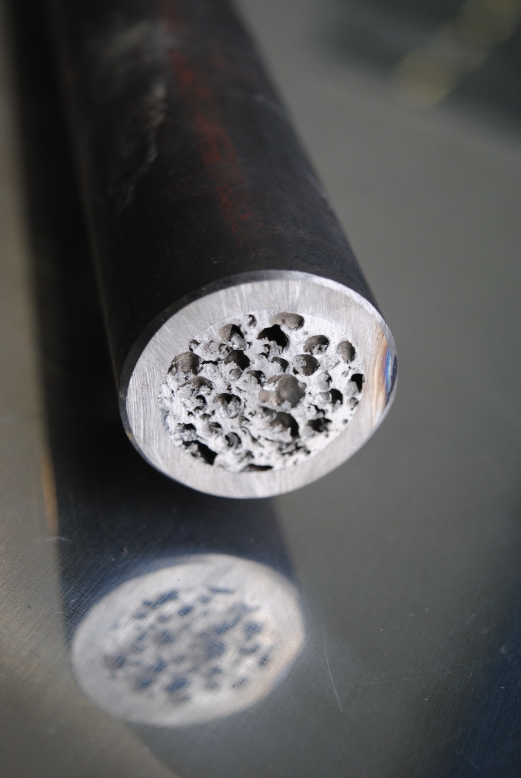 Das angeschnittene Ende eines dunkel beschichteten Rundprofils in Nahaufnahme: In der blank gefrästen Stirnfläche liegt der Aluminiumschaumkern mit deutlich getrennten Poren.