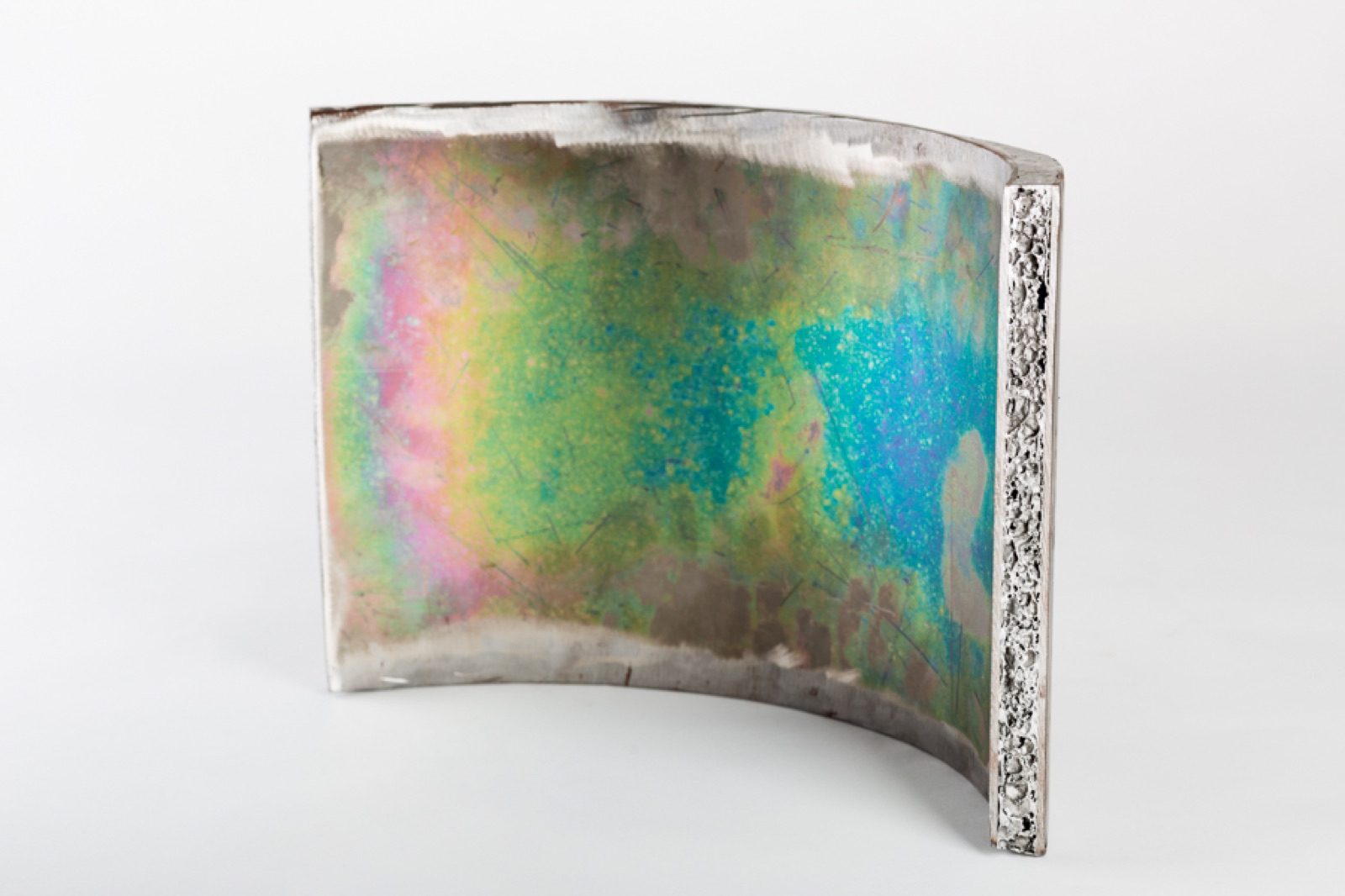Ein gebogenes Sandwichsegment, hochkant stehend; die Außenfläche zeigt Anlauffarben in Gelb, Grün und Blau, an der geschnittenen Stirnseite ist der Aluminiumschaumkern zwischen den Decklagen zu sehen.
