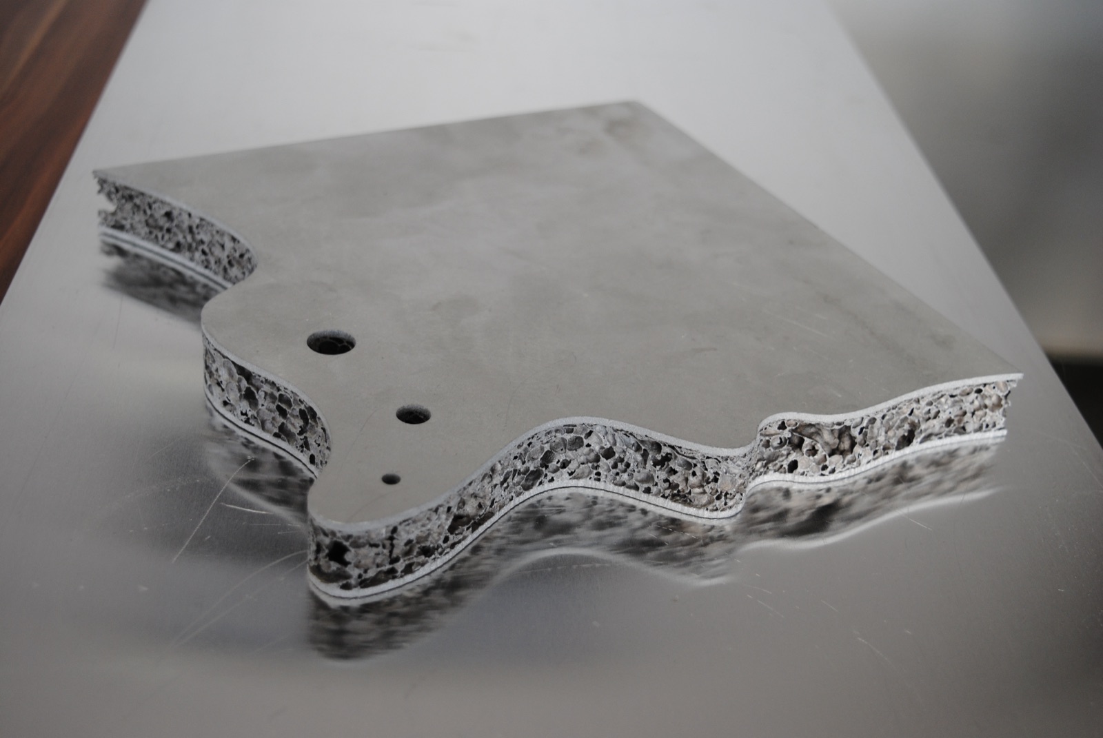 Eine Sandwichplatte mit frei geschnittener, wellenförmiger Außenkontur und drei Bohrungen unterschiedlicher Größe; entlang der gesamten Schnittkante liegt der Aluminiumschaumkern zwischen den beiden Decklagen offen.