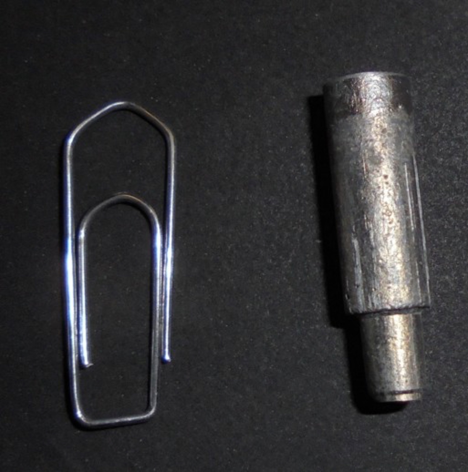 Ein kleiner zylindrischer Stößel aus Aluminiumschaum mit abgesetztem Schaft, daneben eine Büroklammer als Größenvergleich; beide liegen auf dunklem Untergrund.