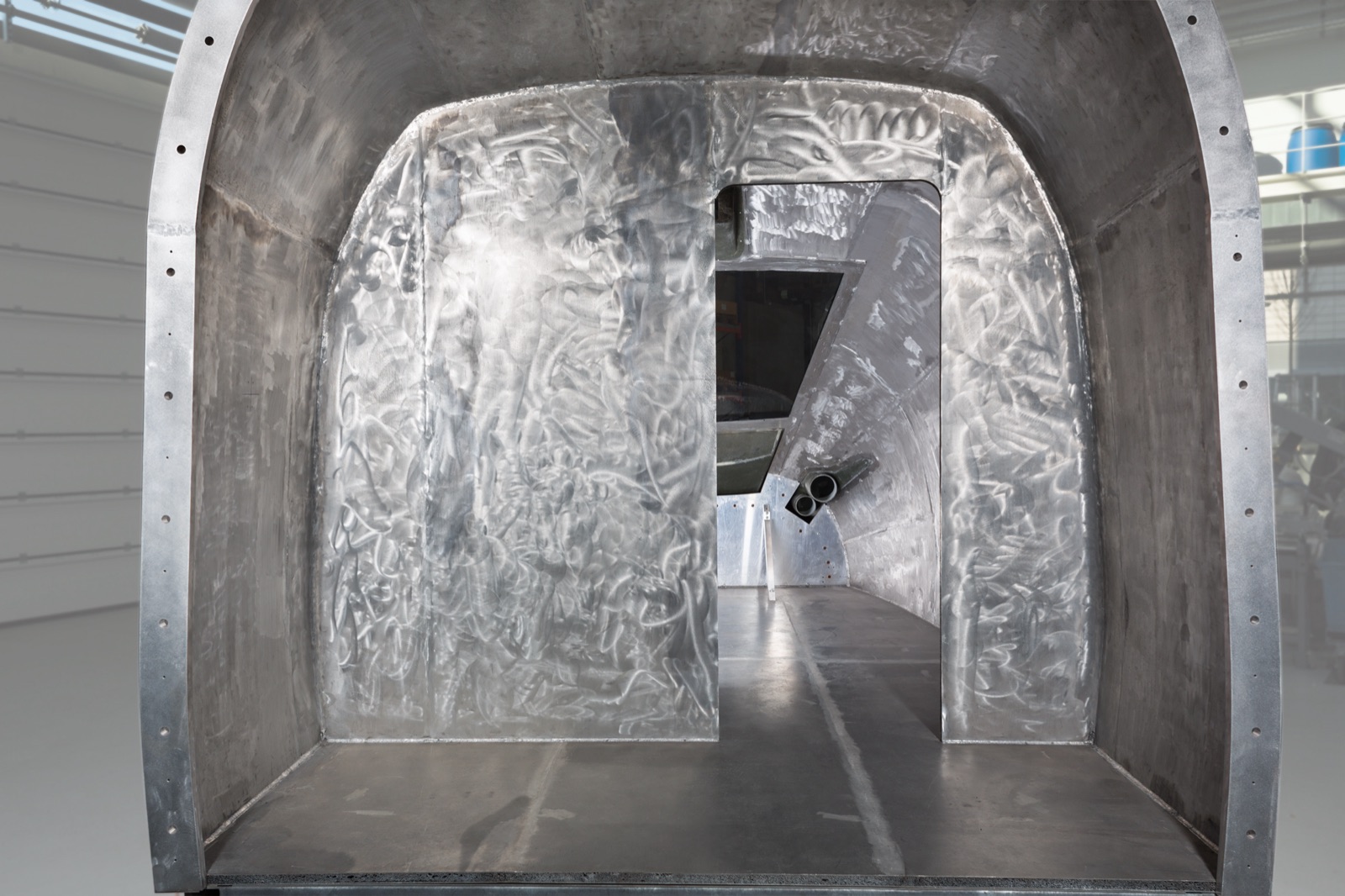 Blick von hinten in eine große, tonnenförmig gewölbte Struktur aus Aluminium-Sandwichplatten: die Triebkopfhaube eines Schnellzugs, mit einem Durchstieg in der Zwischenwand, aufgestellt in einer Fertigungshalle.