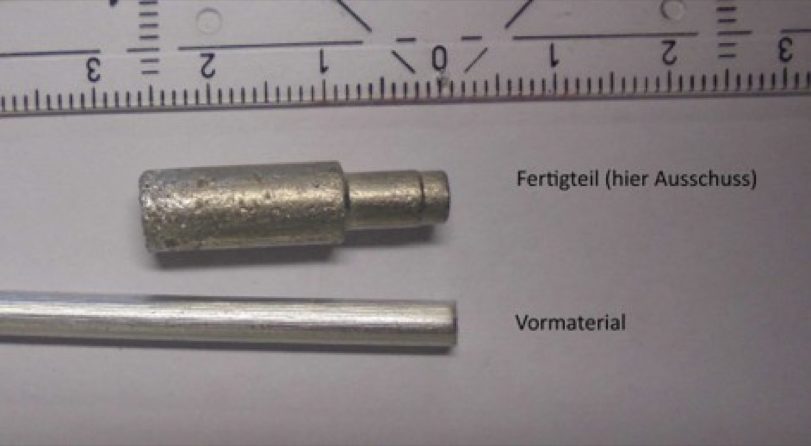 Zwei Teile nebeneinander an einem Lineal mit Zentimeterskala: unten ein glatter Rundstab, beschriftet als Vormaterial, darüber ein abgesetzter Stößel mit aufgeschäumter Oberfläche, beschriftet als Fertigteil und als Ausschussteil gekennzeichnet.